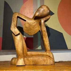 Escultura em madeira "O Pensador" tombada, representando figura humana em pose reflexiva, entalhada artesanalmente com acabamento rústico.