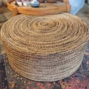Puff redondo grande de fibra natural trançada, em tons de bege e marrom, com acabamento artesanal e formato baixo. Apresentar o Puff de Fibra Natural em detalhe, destacando seu formato, textura, acabamento artesanal e aparência natural para fins de divulgação e venda.