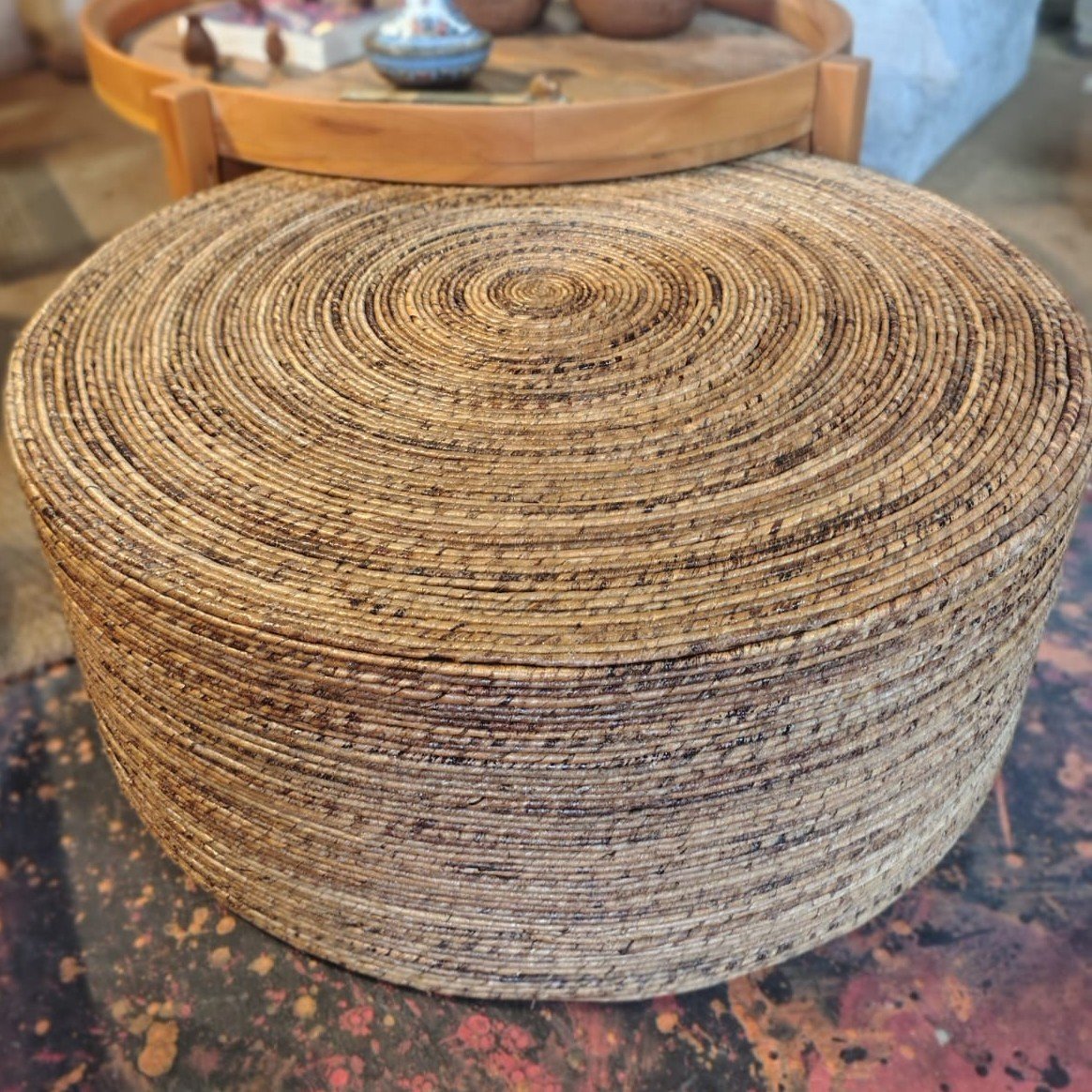 Puff redondo grande de fibra natural trançada, em tons de bege e marrom, com acabamento artesanal e formato baixo. Apresentar o Puff de Fibra Natural em detalhe, destacando seu formato, textura, acabamento artesanal e aparência natural para fins de divulgação e venda.